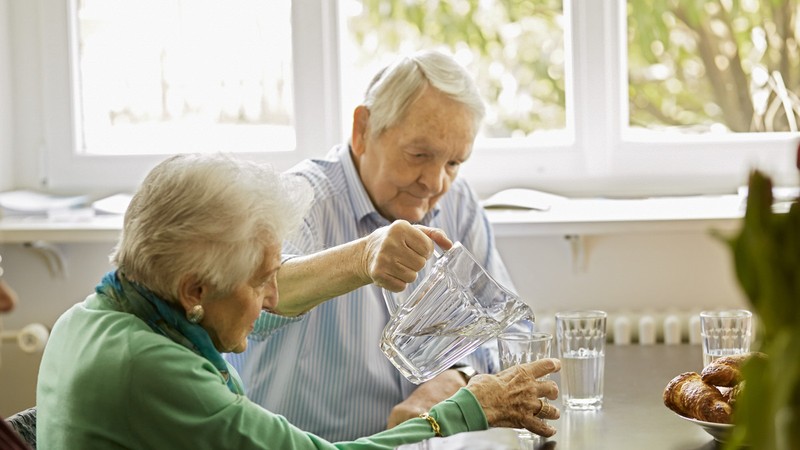 Älterer Mann schenkt älterer Frau ein Glas Wasser ein.